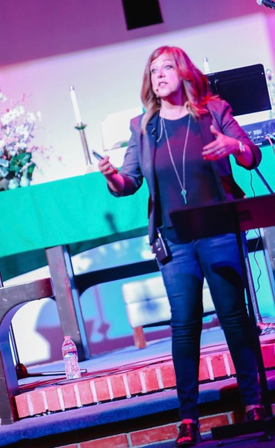 Maria Sainz preaching on stage, sermon notes in one hand, mid-gesture as she teaches a live audience.