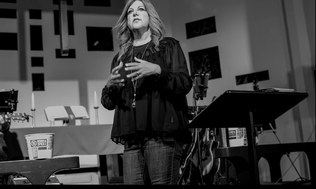 Black-and-white documentary shot of Maria Sainz teaching, microphone in hand, engaged with a room of listeners.
