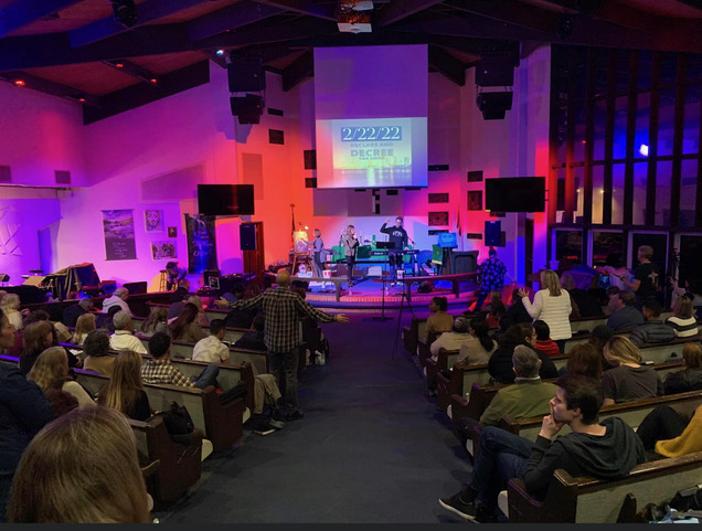 Wide shot of Maria Sainz leading a gathering with a full congregation standing in worship under warm stage lighting.