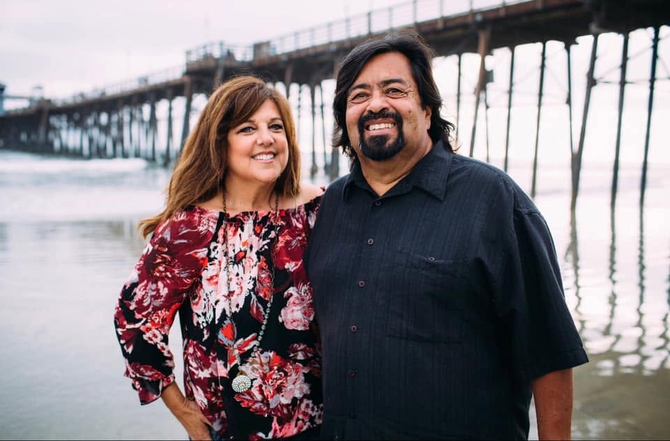 Maria and Rick standing on the beach in front of a wooden pier at Oceanside.
